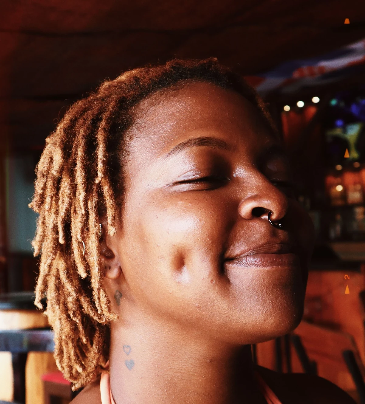 A woman with short textured locs and dimples smiles with her eyes softly closed in warm daylight.