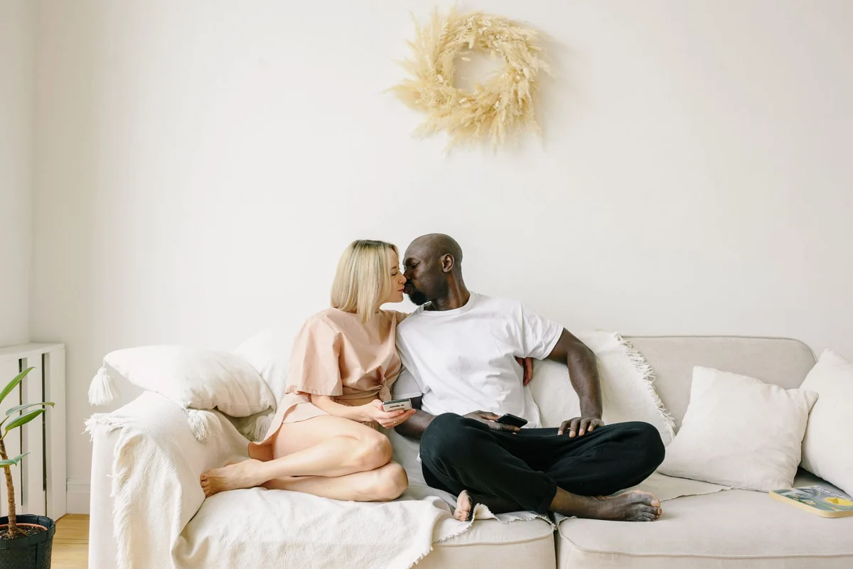 A couple leaning toward each other on a cream sofa in a sunlit room.